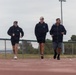 Three individuals are jogging or walking on an outdoor running track, all dressed in dark athletic wear. The setting appears to be a sports facility with a chain-link fence, light poles, and trees visible in the background under an overcast sky. The group appears to be engaged in a casual fitness activity together.