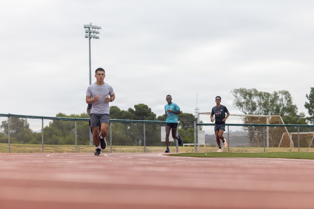 Three male runners are training on a red outdoor athletic track under an overcast sky. The lead runner wears glasses and a gray shirt, followed by two others in teal and dark shirts respectively. The setting appears to be a multi-use sports facility with a soccer goal, chain-link fencing, stadium lights, and trees visible in the background.