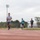 Strength in Every Step: Laps For Life event supports suicide awareness Three male runners are training on a red outdoor athletic track under an overcast sky. The lead runner wears glasses and a gray shirt, followed by two others in teal and dark shirts respectively. The setting appears to be a multi-use sports facility with a soccer goal, chain-link fencing, stadium lights, and trees visible in the background.
