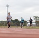 Three male runners are training on a red outdoor athletic track under an overcast sky. The lead runner wears glasses and a gray shirt, followed by two others in teal and dark shirts respectively. The setting appears to be a multi-use sports facility with a soccer goal, chain-link fencing, stadium lights, and trees visible in the background.
