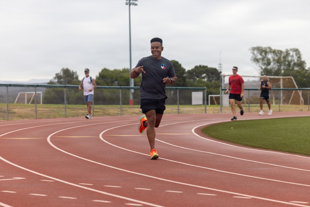 A young man is sprinting on a red outdoor running track, smiling broadly as he rounds a curve. Several other individuals are visible in the background, also using the track and surrounding athletic facility. The setting appears to be a multi-sport complex with soccer goals, floodlights, and green turf infield visible.