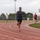 Strength in Every Step: Laps For Life event supports suicide awareness A young man is sprinting on a red outdoor running track, smiling broadly as he rounds a curve. Several other individuals are visible in the background, also using the track and surrounding athletic facility. The setting appears to be a multi-sport complex with soccer goals, floodlights, and green turf infield visible.