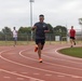 A young man is sprinting on a red outdoor running track, smiling broadly as he rounds a curve. Several other individuals are visible in the background, also using the track and surrounding athletic facility. The setting appears to be a multi-sport complex with soccer goals, floodlights, and green turf infield visible.