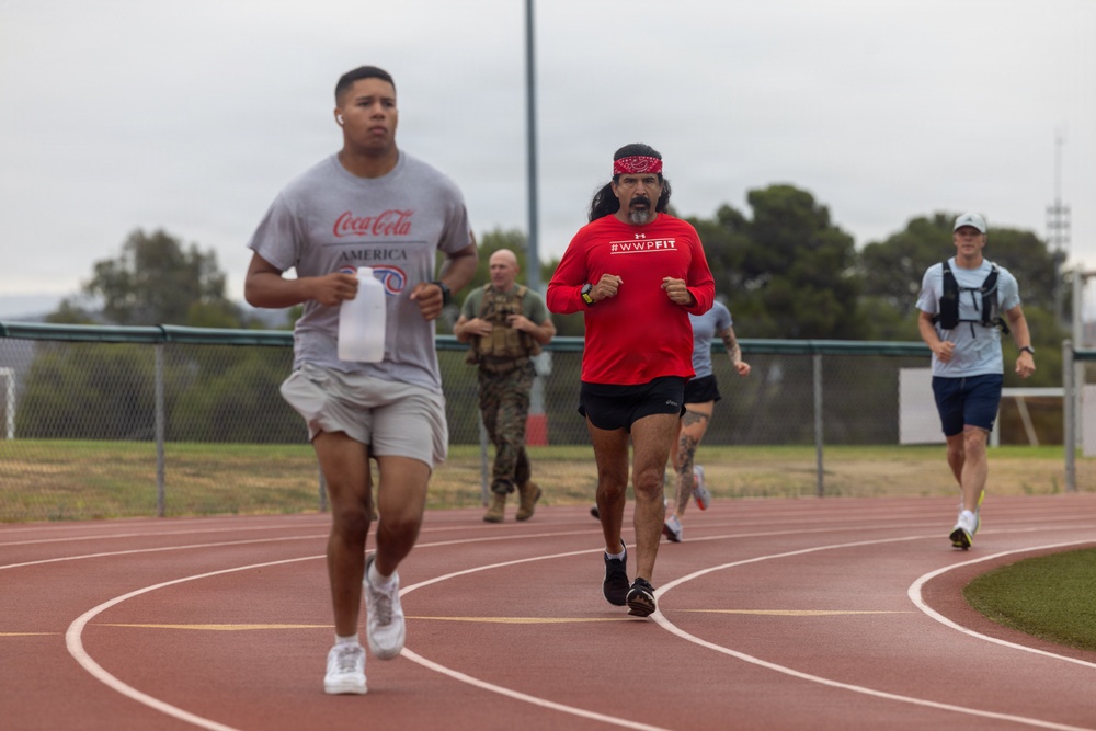 A group of people are running on a red outdoor athletic track under an overcast sky. The runners include individuals in casual athletic wear, with one wearing a Coca-Cola America t-shirt and another in a red Under Armour #WWPFIT shirt with a bandana headband. In the background, a person in military camouflage uniform is also participating in the run.