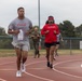 A group of people are running on a red outdoor athletic track under an overcast sky. The runners include individuals in casual athletic wear, with one wearing a Coca-Cola America t-shirt and another in a red Under Armour #WWPFIT shirt with a bandana headband. In the background, a person in military camouflage uniform is also participating in the run.