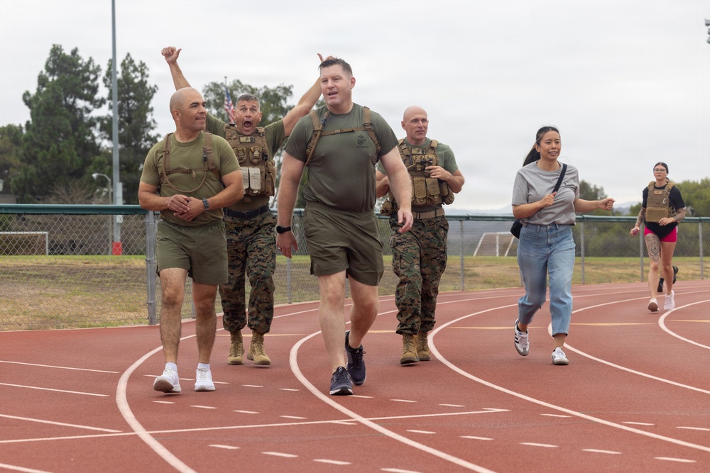 A group of individuals are walking or jogging on a red outdoor athletic track, appearing to be participating in a fitness or ruck march event. Several participants are wearing U.S. Marine Corps uniforms with tactical vests/plate carriers, while others are in civilian athletic attire. The atmosphere appears celebratory or energetic, with at least one person raising a fist in the air.