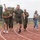 Strength in Every Step: Laps For Life event supports suicide awareness A group of individuals are walking or jogging on a red outdoor athletic track, appearing to be participating in a fitness or ruck march event. Several participants are wearing U.S. Marine Corps uniforms with tactical vests/plate carriers, while others are in civilian athletic attire. The atmosphere appears celebratory or energetic, with at least one person raising a fist in the air.