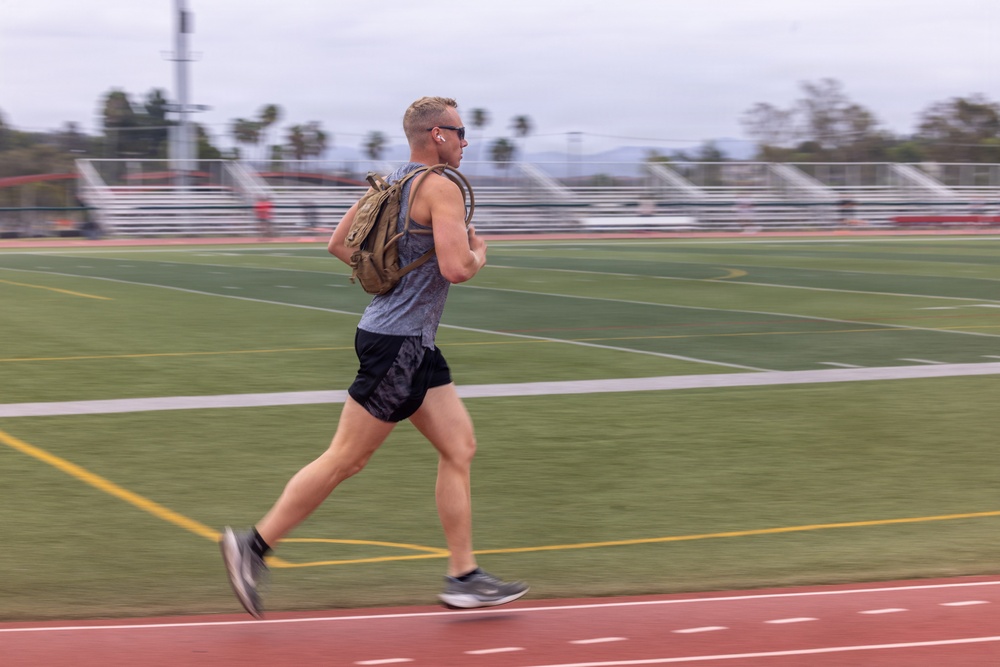 A male athlete is running on a red outdoor track while wearing a backpack, sunglasses, and earbuds. The background shows a green football field with yard lines, bleachers, light poles, palm trees, and mountains under an overcast sky. Motion blur on the subject and background conveys a sense of speed.