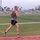 Strength in Every Step: Laps For Life event supports suicide awareness A male athlete is running on a red outdoor track while wearing a backpack, sunglasses, and earbuds. The background shows a green football field with yard lines, bleachers, light poles, palm trees, and mountains under an overcast sky. Motion blur on the subject and background conveys a sense of speed.