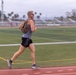 A male athlete is running on a red outdoor track while wearing a backpack, sunglasses, and earbuds. The background shows a green football field with yard lines, bleachers, light poles, palm trees, and mountains under an overcast sky. Motion blur on the subject and background conveys a sense of speed.