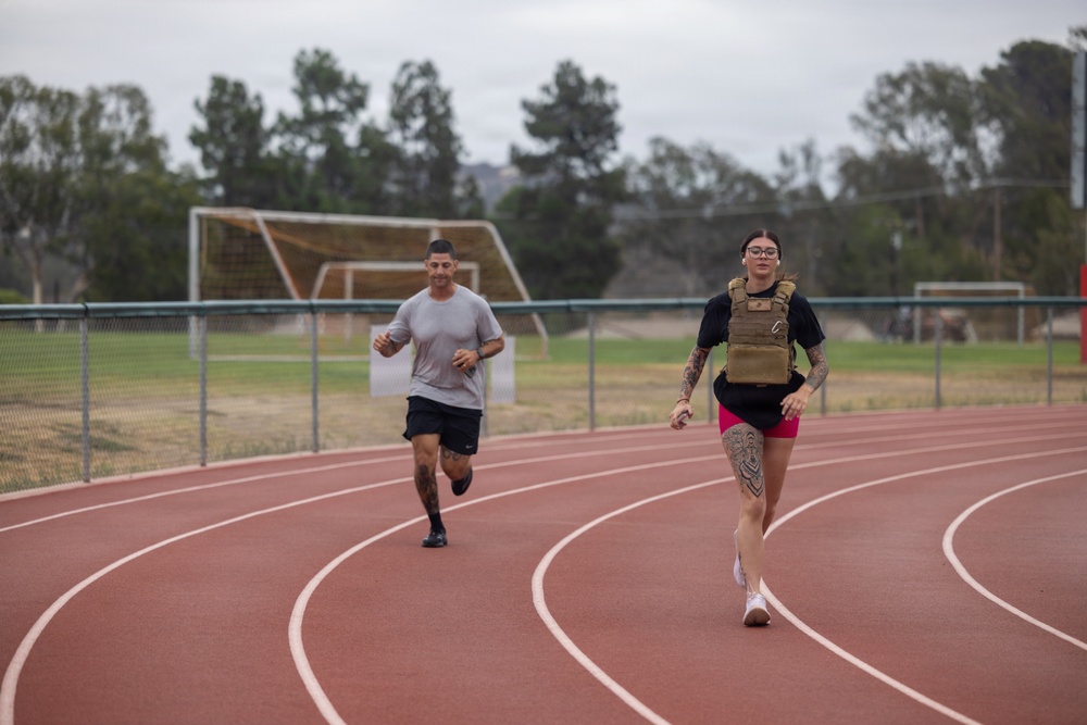 Two individuals are running on a red outdoor athletic track, with a soccer field visible in the background. The woman in the foreground is wearing a tactical/weighted vest over a black t-shirt and pink shorts, while the man behind her wears a gray t-shirt and black shorts. Both have visible tattoos and appear to be engaged in a fitness training session.