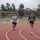 Strength in Every Step: Laps For Life event supports suicide awareness Two individuals are running on a red outdoor athletic track, with a soccer field visible in the background. The woman in the foreground is wearing a tactical/weighted vest over a black t-shirt and pink shorts, while the man behind her wears a gray t-shirt and black shorts. Both have visible tattoos and appear to be engaged in a fitness training session.