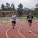 Two individuals are running on a red outdoor athletic track, with a soccer field visible in the background. The woman in the foreground is wearing a tactical/weighted vest over a black t-shirt and pink shorts, while the man behind her wears a gray t-shirt and black shorts. Both have visible tattoos and appear to be engaged in a fitness training session.