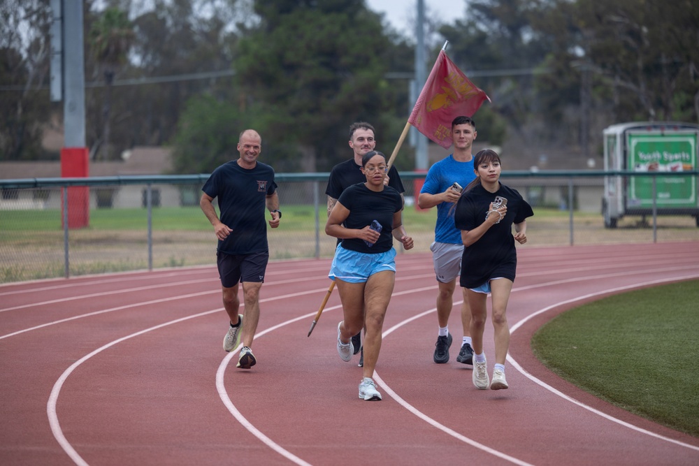 A group of five individuals are running together on a red athletic track, with one person carrying what appears to be a red military-style flag on a wooden pole. The group appears to be participating in an organized run or relay event, likely on a military or school facility. The setting includes a well-maintained track, green infield grass, and trees in the background.