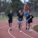 A group of five individuals are running together on a red athletic track, with one person carrying what appears to be a red military-style flag on a wooden pole. The group appears to be participating in an organized run or relay event, likely on a military or school facility. The setting includes a well-maintained track, green infield grass, and trees in the background.