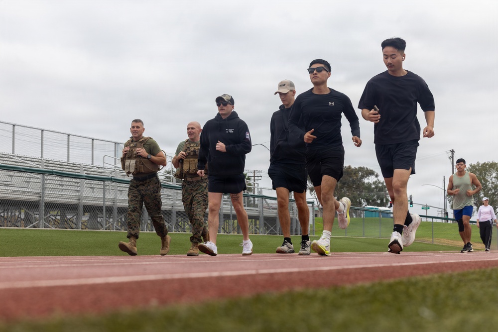 A group of individuals are running or jogging on an outdoor athletic track, with two people in military camouflage uniforms and tactical vests accompanying civilians in casual athletic wear. The setting appears to be a military base or public sports facility with bleachers visible in the background. The overcast sky and green grass infield suggest a temperate coastal environment.