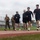 Strength in Every Step: Laps For Life event supports suicide awareness A group of individuals are running or jogging on an outdoor athletic track, with two people in military camouflage uniforms and tactical vests accompanying civilians in casual athletic wear. The setting appears to be a military base or public sports facility with bleachers visible in the background. The overcast sky and green grass infield suggest a temperate coastal environment.