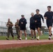 A group of individuals are running or jogging on an outdoor athletic track, with two people in military camouflage uniforms and tactical vests accompanying civilians in casual athletic wear. The setting appears to be a military base or public sports facility with bleachers visible in the background. The overcast sky and green grass infield suggest a temperate coastal environment.