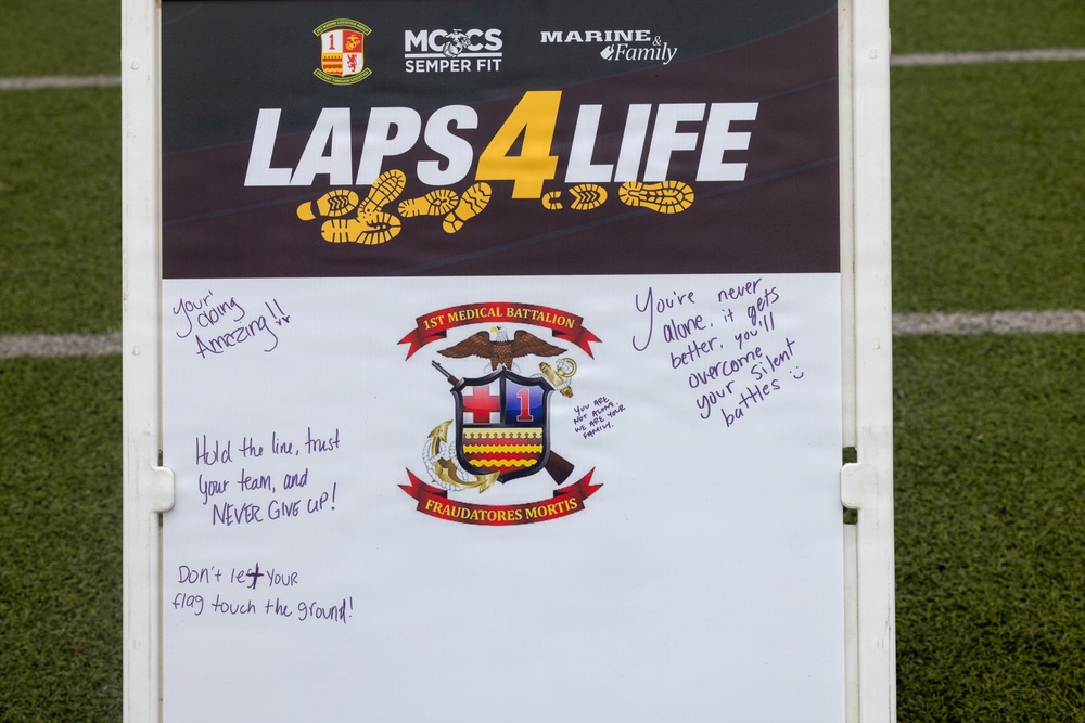 A sign for a 'Laps 4 Life' event organized by MCCS Semper Fit and Marine &amp; Family programs, associated with the 1st Medical Battalion. The whiteboard portion contains handwritten motivational messages supporting mental health and perseverance. The event appears to be a fitness/awareness fundraiser held on a grass athletic field.