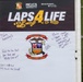 A sign for a 'Laps 4 Life' event organized by MCCS Semper Fit and Marine &amp; Family programs, associated with the 1st Medical Battalion. The whiteboard portion contains handwritten motivational messages supporting mental health and perseverance. The event appears to be a fitness/awareness fundraiser held on a grass athletic field.