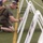Strength in Every Step: Laps For Life event supports suicide awareness A military service member in olive drab PT (physical training) gear and a tactical backpack kneels on an artificial turf athletic field, writing or signing on a display board mounted on a white easel. Several similar easels with photographs or memorial displays are arranged in a row on the field. Other personnel in military camouflage uniforms are visible in the background, suggesting a formal or ceremonial event.