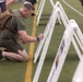 A military service member in olive drab PT (physical training) gear and a tactical backpack kneels on an artificial turf athletic field, writing or signing on a display board mounted on a white easel. Several similar easels with photographs or memorial displays are arranged in a row on the field. Other personnel in military camouflage uniforms are visible in the background, suggesting a formal or ceremonial event.