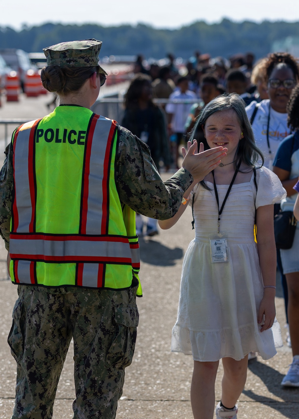 A military police officer in U.S. Navy digital camouflage uniform and high-visibility safety vest interacts with a young girl wearing a white dress and lanyard badge at what appears to be a public airshow or military open house event. The officer appears to be giving the smiling girl a high-five. A crowd of attendees is visible in the background on what appears to be a tarmac or airfield.