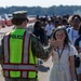 A military police officer in U.S. Navy digital camouflage uniform and high-visibility safety vest interacts with a young girl wearing a white dress and lanyard badge at what appears to be a public airshow or military open house event. The officer appears to be giving the smiling girl a high-five. A crowd of attendees is visible in the background on what appears to be a tarmac or airfield.