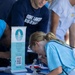 A young blonde female student in a light blue shirt is looking through a microscope at a table display booth for the Math and Science Academy at Clear Lakes High School. The booth features a pull-up banner with a QR code and the text 'Riding waves of success since 1996.' Several other individuals in the background, including someone wearing a 'Clear Lakes Volleyball' shirt, are gathered around the table.