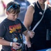 A young boy wearing a Shadow the Hedgehog t-shirt and blue sunglasses on his head is observing a spider being placed on his arm by an adult woman in a black sleeveless top. The setting appears to be an outdoor public event or fair, with a blurred crowd visible in the background. The boy looks focused and cautious while examining the spider.