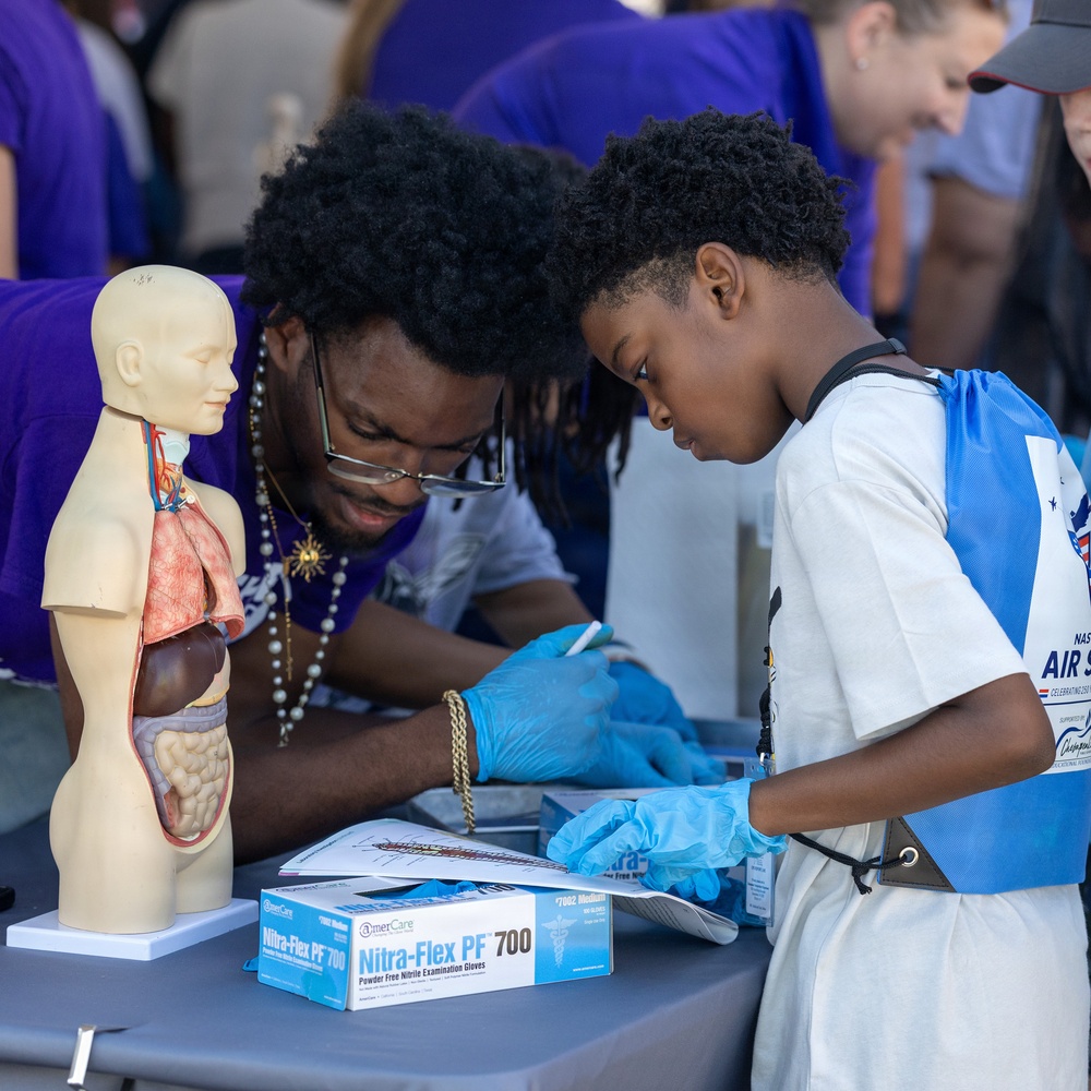 A young child wearing blue nitrile gloves and a NASA Air Show vest is engaged in a hands-on medical/science activity at an educational booth, guided by an adult volunteer in a purple shirt. The table features an anatomical human torso model with visible organs, a box of Nitra-Flex PF 700 nitrile examination gloves, and printed worksheets or activity materials. The setting appears to be an outdoor science or STEM education event.