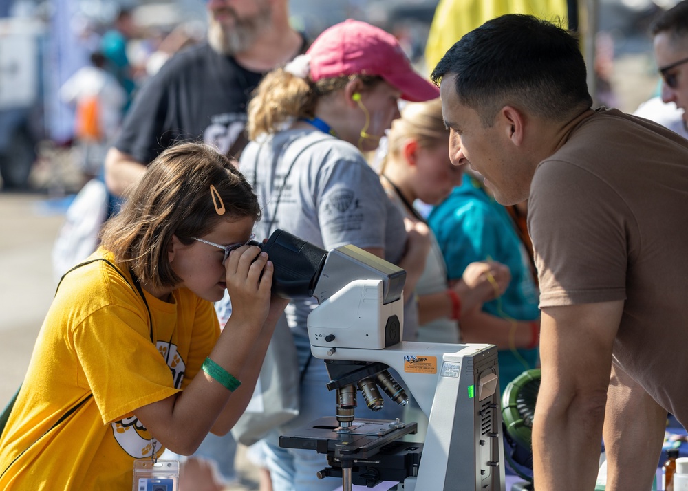 A young girl in a yellow t-shirt looks through a compound microscope at what appears to be an outdoor science fair or educational event. An adult man in a brown shirt leans in attentively, likely explaining or guiding her observation. The background shows a crowd of other participants engaged in similar activities at various booths or stations.
