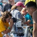 A young girl in a yellow t-shirt looks through a compound microscope at what appears to be an outdoor science fair or educational event. An adult man in a brown shirt leans in attentively, likely explaining or guiding her observation. The background shows a crowd of other participants engaged in similar activities at various booths or stations.