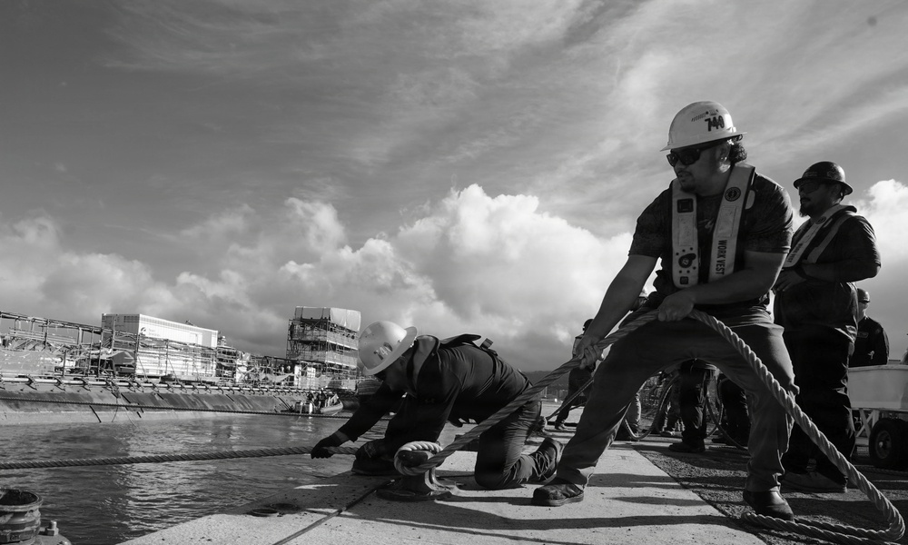 A black and white photograph showing workers in hard hats and safety vests hauling a thick mooring rope on what appears to be a dock or vessel deck. The foreground worker is braced in a low stance pulling the rope with significant effort, while a second worker crouches near the water's edge. An industrial shipyard or construction facility is visible in the background.