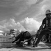 A black and white photograph showing workers in hard hats and safety vests hauling a thick mooring rope on what appears to be a dock or vessel deck. The foreground worker is braced in a low stance pulling the rope with significant effort, while a second worker crouches near the water's edge. An industrial shipyard or construction facility is visible in the background.