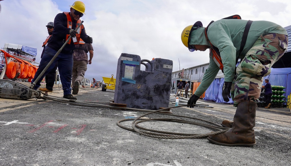 Workers are engaged in heavy rigging or cable-handling operations at what appears to be a port or industrial construction site. One worker in military camouflage uniform and a yellow hard hat is bending down to manage a coiled steel cable, while another worker in an orange safety vest uses a long-handled tool nearby. The site contains various equipment, barriers, and temporary structures in the background.