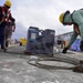 Workers are engaged in heavy rigging or cable-handling operations at what appears to be a port or industrial construction site. One worker in military camouflage uniform and a yellow hard hat is bending down to manage a coiled steel cable, while another worker in an orange safety vest uses a long-handled tool nearby. The site contains various equipment, barriers, and temporary structures in the background.