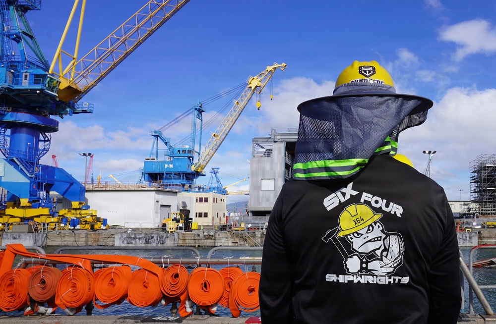 A worker wearing a black jacket labeled 'Six Four Shipwrights' with 'X64' branding stands at a shipyard or dry dock facility, viewed from behind. The worker is equipped with a yellow hard hat fitted with a mesh sun/insect shield and is overlooking an industrial waterfront area with large blue and yellow cranes. Multiple coiled orange fire hoses are visible in the foreground along the dock edge.