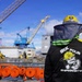 A worker wearing a black jacket labeled 'Six Four Shipwrights' with 'X64' branding stands at a shipyard or dry dock facility, viewed from behind. The worker is equipped with a yellow hard hat fitted with a mesh sun/insect shield and is overlooking an industrial waterfront area with large blue and yellow cranes. Multiple coiled orange fire hoses are visible in the foreground along the dock edge.