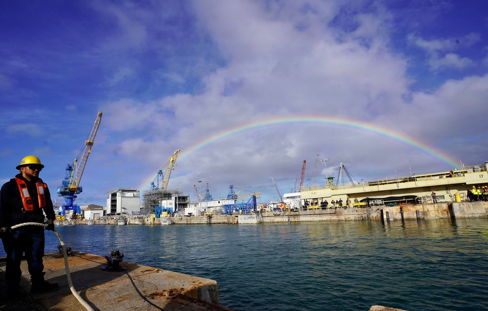 A naval shipyard or industrial port facility is shown with multiple cranes and dry docks visible along the waterfront. A full rainbow arcs across a partly cloudy blue sky above the facility. In the foreground, a worker wearing a yellow hard hat, safety glasses, and an orange life vest stands on a dock or vessel near mooring lines.