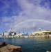A naval shipyard or industrial port facility is shown with multiple cranes and dry docks visible along the waterfront. A full rainbow arcs across a partly cloudy blue sky above the facility. In the foreground, a worker wearing a yellow hard hat, safety glasses, and an orange life vest stands on a dock or vessel near mooring lines.