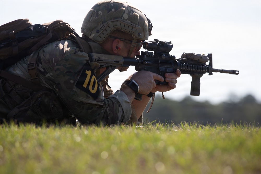 A soldier in full combat gear is in a prone firing position on a grass field, aiming a rifle equipped with an optical scope. The individual wears multicam camouflage, a tactical helmet, and displays a yellow number '10' identifier on their arm, suggesting a training exercise or competition. The image is taken from a low angle, emphasizing the shooter's position and weapon.