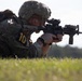A soldier in full combat gear is in a prone firing position on a grass field, aiming a rifle equipped with an optical scope. The individual wears multicam camouflage, a tactical helmet, and displays a yellow number '10' identifier on their arm, suggesting a training exercise or competition. The image is taken from a low angle, emphasizing the shooter's position and weapon.
