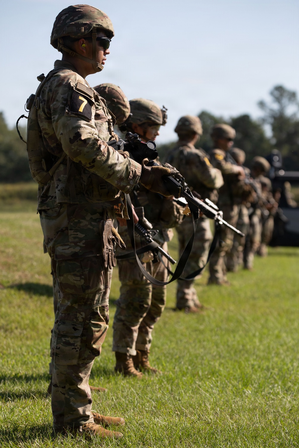 A line of military soldiers in full combat gear stands at attention on a grassy field, each holding rifles. The foreground soldier prominently displays a yellow number '7' patch on their left arm, wearing OCP (Operational Camouflage Pattern) uniforms with helmets and tactical equipment. The formation appears to be part of a training exercise, demonstration, or formal military event.