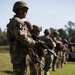 A line of military soldiers in full combat gear stands at attention on a grassy field, each holding rifles. The foreground soldier prominently displays a yellow number '7' patch on their left arm, wearing OCP (Operational Camouflage Pattern) uniforms with helmets and tactical equipment. The formation appears to be part of a training exercise, demonstration, or formal military event.