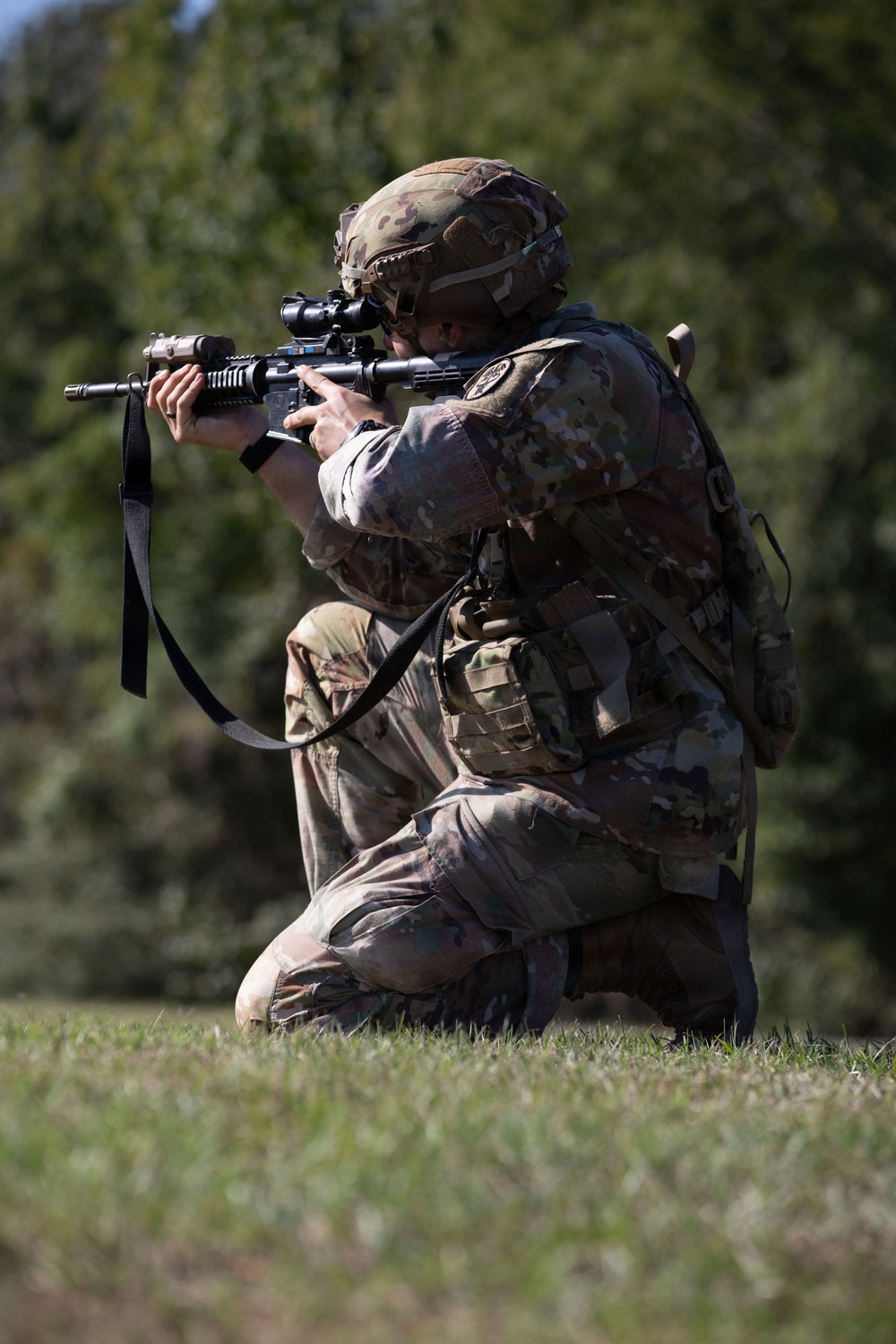 A soldier in full combat gear is kneeling on grass in a firing position, aiming a rifle equipped with an optic scope and accessories. The individual is wearing Operational Camouflage Pattern (OCP) uniform, tactical helmet, and a plate carrier vest with pouches. The setting appears to be an outdoor training or exercise environment with trees visible in the background.