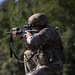 A soldier in full combat gear is kneeling on grass in a firing position, aiming a rifle equipped with an optic scope and accessories. The individual is wearing Operational Camouflage Pattern (OCP) uniform, tactical helmet, and a plate carrier vest with pouches. The setting appears to be an outdoor training or exercise environment with trees visible in the background.