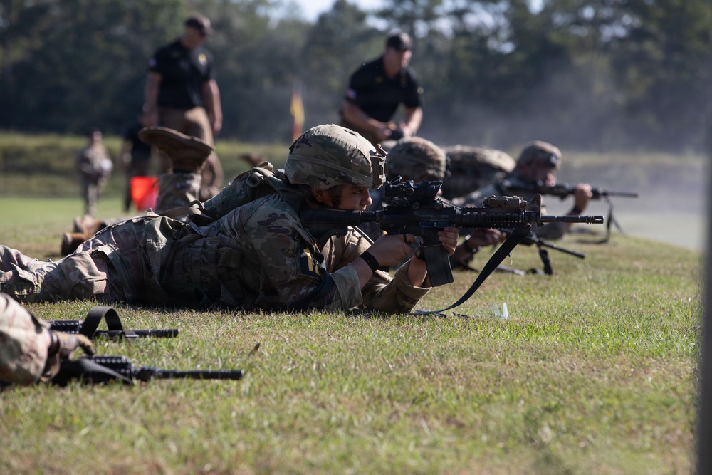Military personnel in camouflage uniforms are conducting a prone firing exercise on a grass range. Multiple soldiers are lying in the prone position aiming rifles downrange, while supervisors in black uniforms observe from a standing position in the background. The scene appears to be a structured marksmanship qualification or training event.