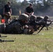 Military personnel in camouflage uniforms are conducting a prone firing exercise on a grass range. Multiple soldiers are lying in the prone position aiming rifles downrange, while supervisors in black uniforms observe from a standing position in the background. The scene appears to be a structured marksmanship qualification or training event.
