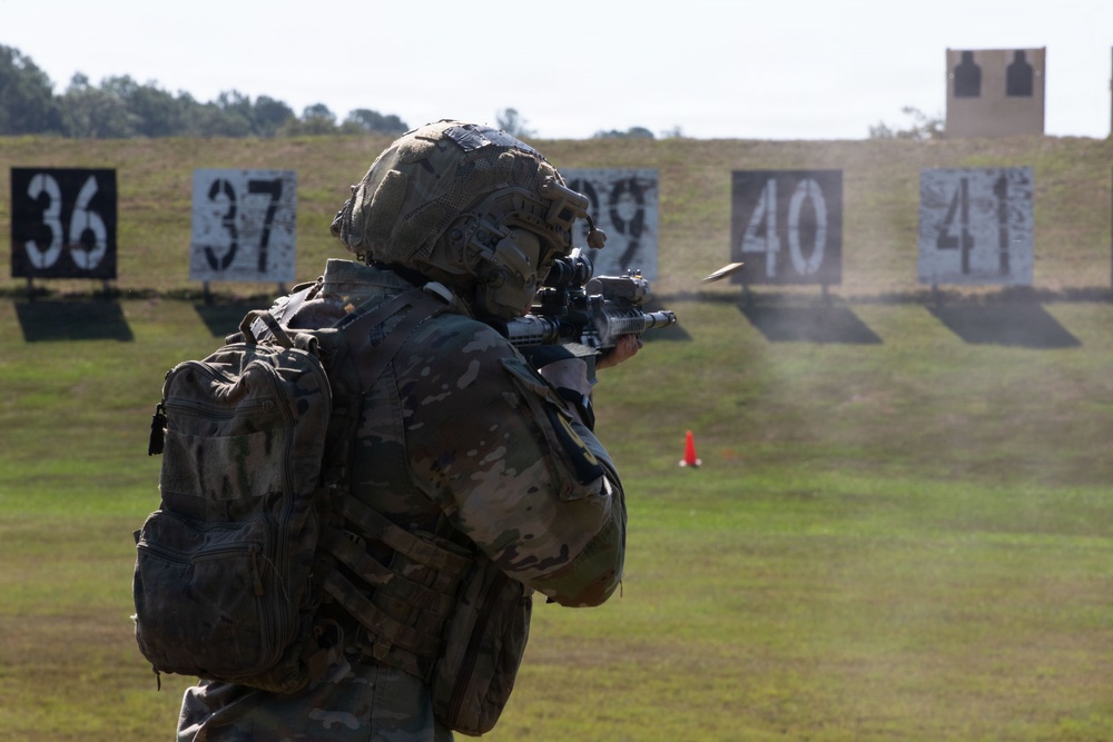 A soldier in full combat gear, including a tactical helmet and multicam camouflage uniform with a backpack, is firing a rifle on a military shooting range. Numbered target boards (36, 37, 39, 40, 41) are visible downrange on a grassy field. A spent casing appears to be ejecting from the weapon, captured mid-flight.